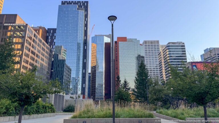 Landscaped downtown Calgary park with trees, grasses, pathway, lamp post, and modern office towers surrounding it.