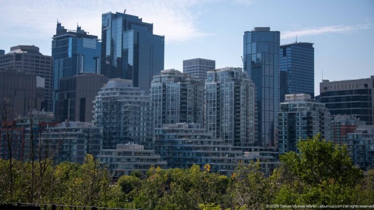 Dense Calgary skyline of residential balcony towers and taller glass office buildings rising beyond green trees.
