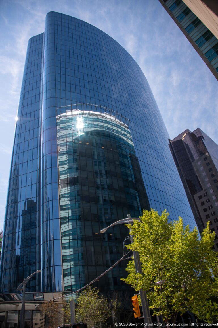 Curved reflective blue-glass office tower rising above trees and traffic lights in downtown Calgary.