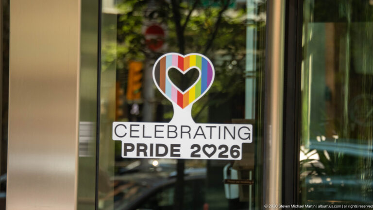 Rainbow heart window decal above the words Celebrating Pride 2026, with trees and street reflections in the glass.