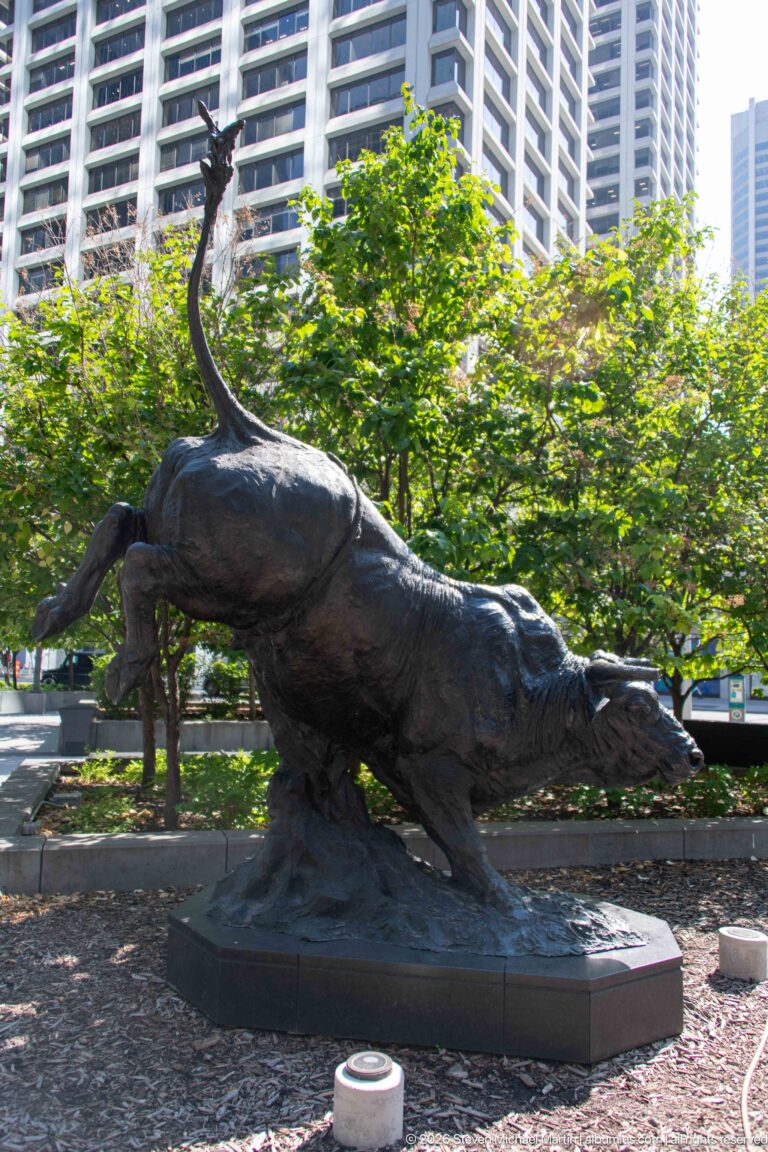 Large bronze bull sculpture lunging forward on a low plinth among trees and downtown residential towers.