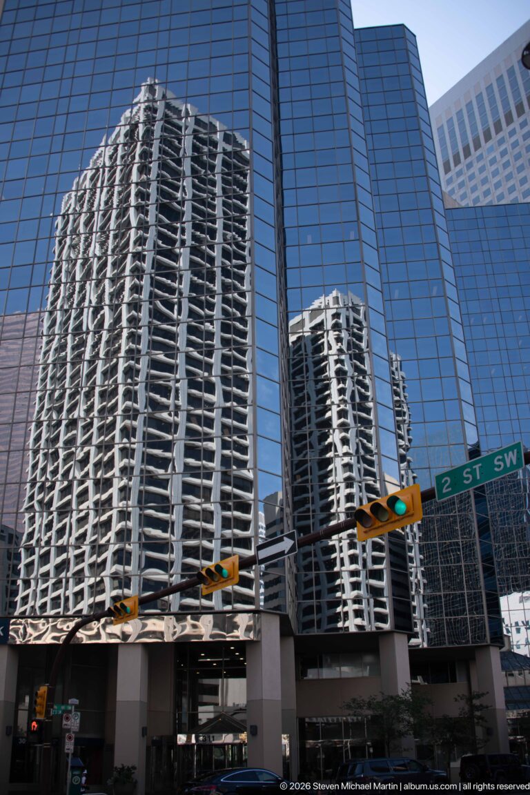 Residential towers reflected in a blue-glass office building above traffic lights and a 2 Street SW sign in Calgary.