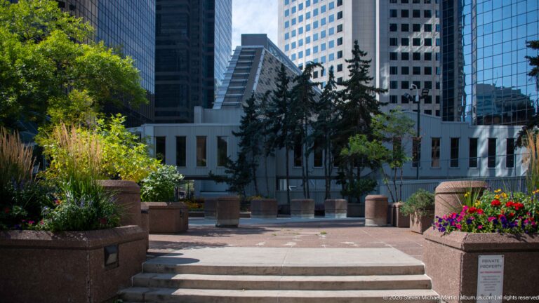 Landscaped downtown Calgary plaza with steps, raised flower beds, trees, and office towers surrounding a low building.