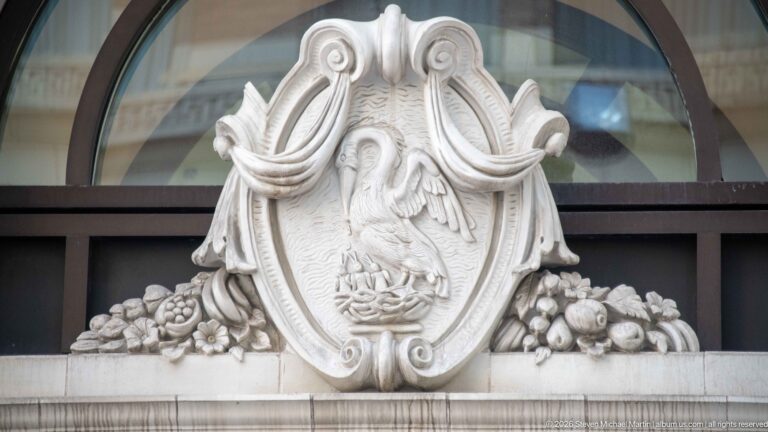 Carved stone crest with a pelican-like bird, scrollwork, flowers, and fruit beneath an arched window.
