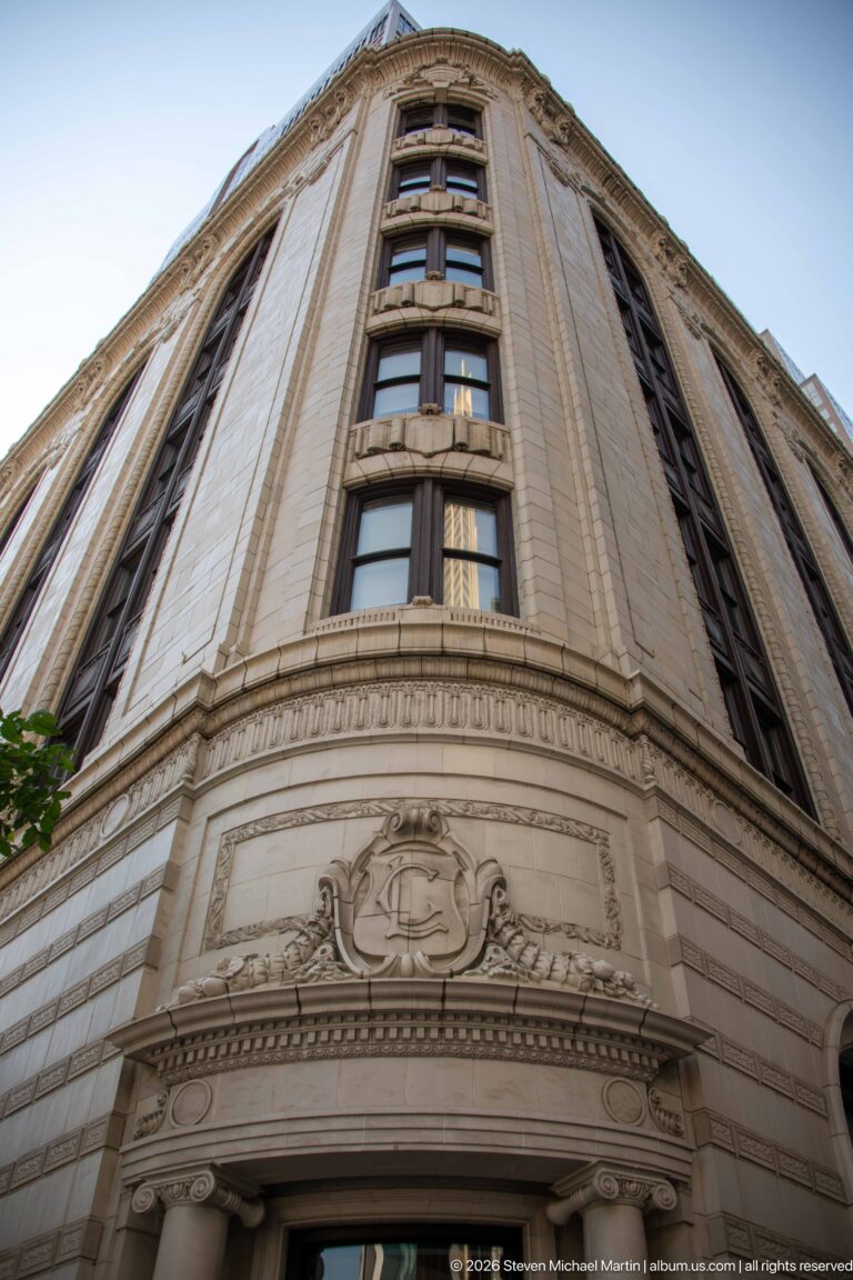 Low-angle view up an ornate pale masonry corner building with vertical windows, dark panels, and carved decoration.