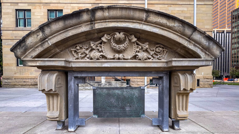 Freestanding carved stone arch with floral ornament and curved pediment framing a bronze courthouse history plaque.