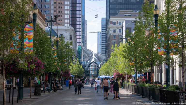 Tree-lined Stephen Avenue with pedestrians, hanging flowers, colorful banners, patios, historic façades, and towers beyond.
