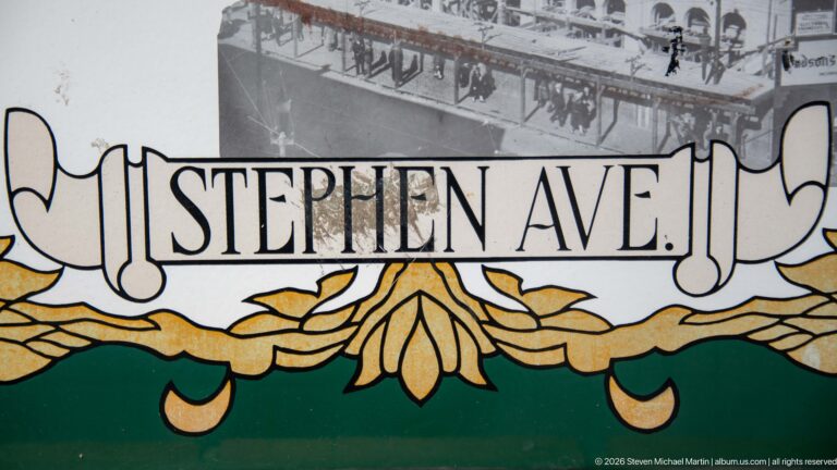 Decorative Stephen Ave. sign with black lettering, gold floral ornament, green border, and historic street photograph behind.