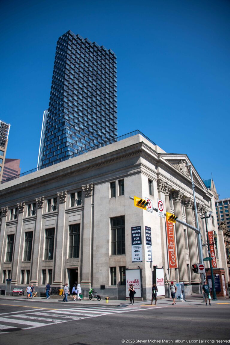 Historic Bank of Montreal stone building at a Calgary intersection with a modern residential tower rising behind.