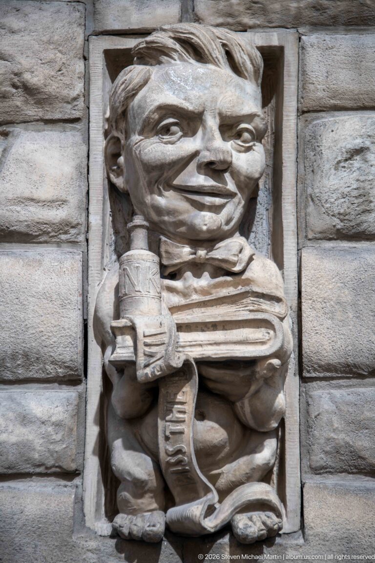 Carved stone caricature of a seated figure holding an object beside stacked papers or ledgers in a masonry wall.