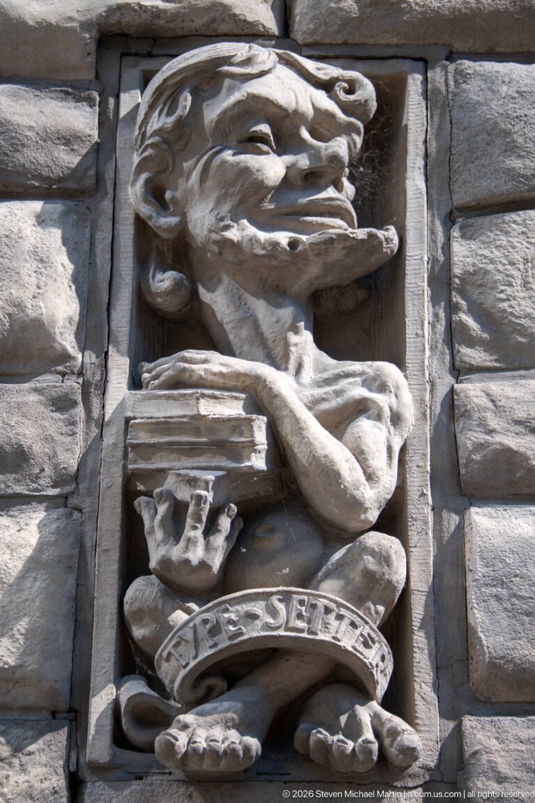 Stone architectural relief labeled The Spender, showing a caricatured figure with stacked objects and swirling forms.