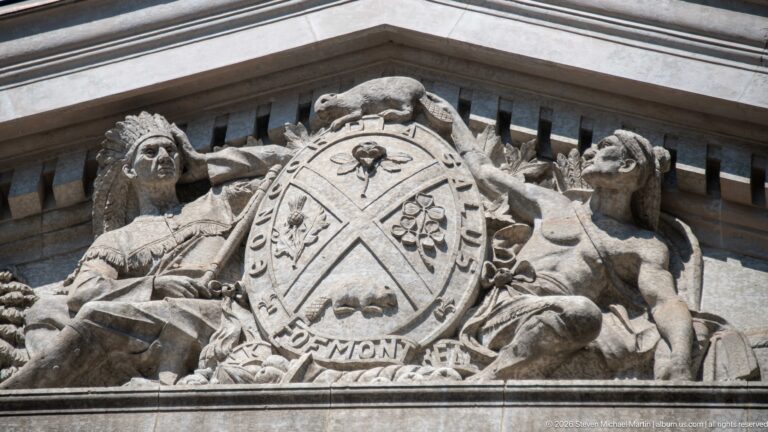 Carved stone pediment relief with allegorical figures, animals, foliage, and central shield on the Bank of Montreal building.