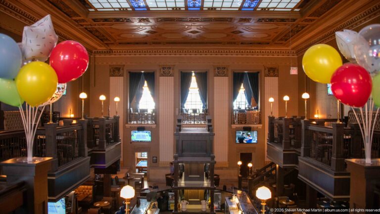 Grand Bank & Baron interior with coffered ceiling, tall windows, dark galleries, globe lights, and colorful balloons.