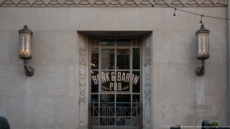 Bank & Baron P.U.B. window framed by carved stone details and two tall wall lanterns.