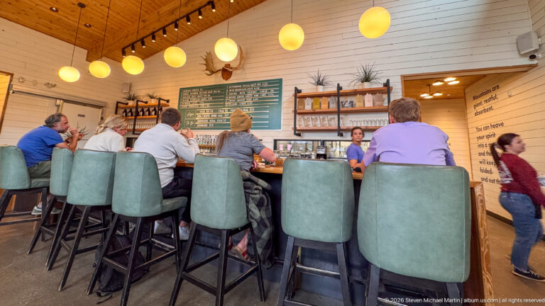 Wide taproom interior with patrons on tall green stools at a wood bar beneath globe lights and a large menu board.
