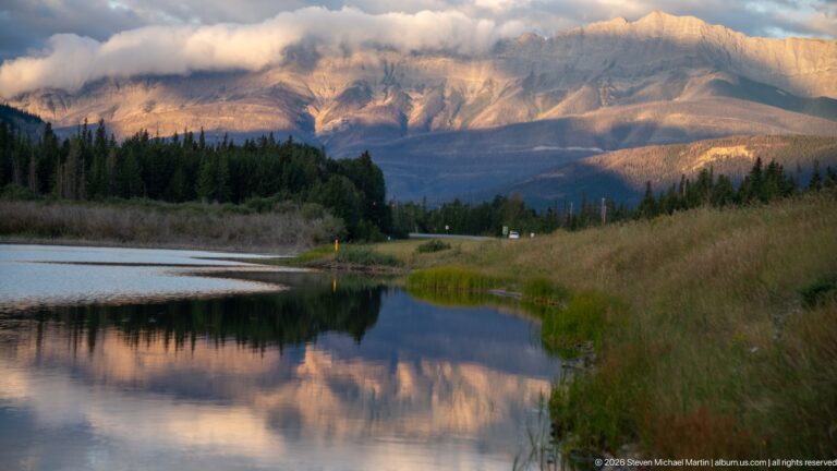 Sunlit mountain faces beneath clouds beyond reflective water, grasses, conifers, and a roadway.