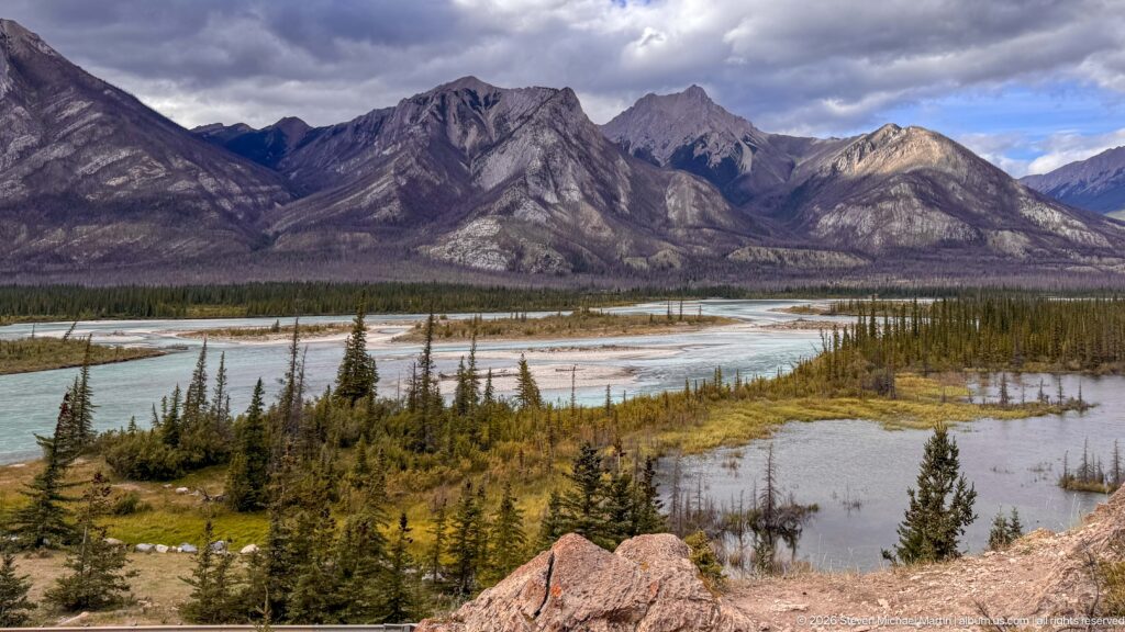 Braided pale-blue river channels, gravel bars, and forested islands beneath steep gray mountain slopes.