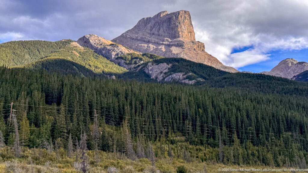 Flat-topped sedimentary mountain with steep cliffs above conifer forest and foreground power lines.