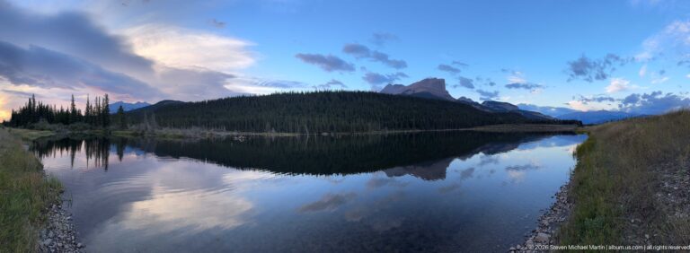 Calm lake reflecting a forested ridge, distant mountain peaks, and blue and pink twilight clouds.
