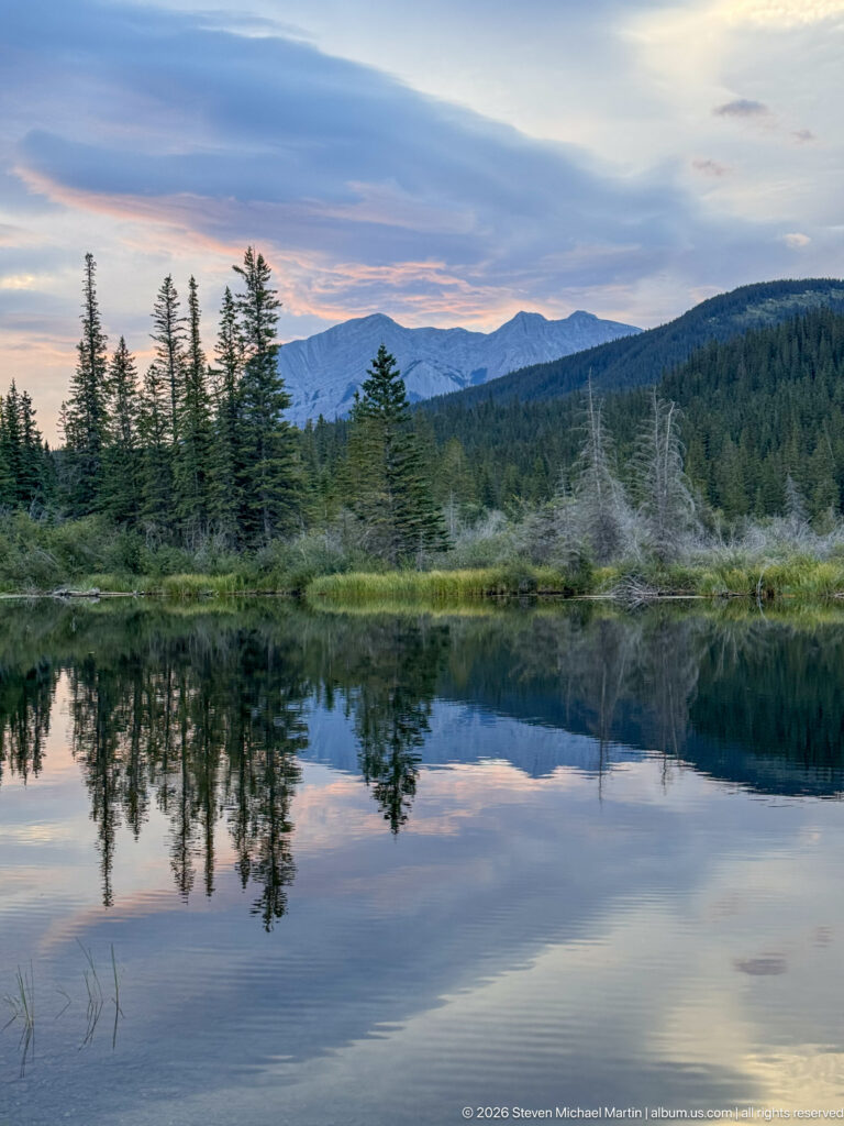 Still pond reflecting conifers, marsh vegetation, evening clouds, and a long mountain ridge.