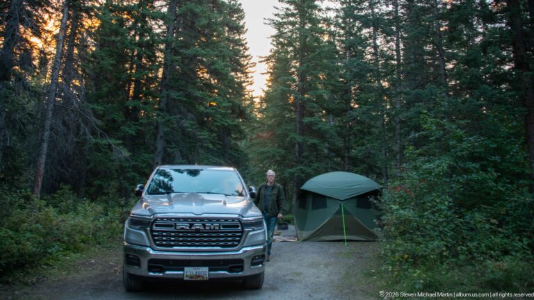 Pickup truck, green tent, and person at a compact campsite surrounded by dense conifer forest.