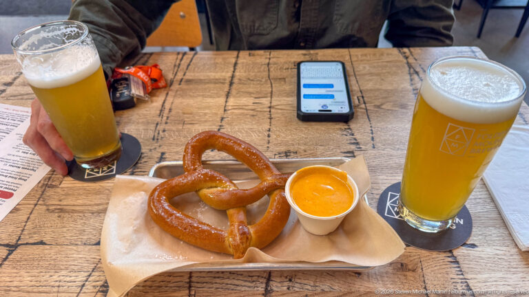 Salted pretzel with mustard between two tall pale draft beers on a rustic wood table.