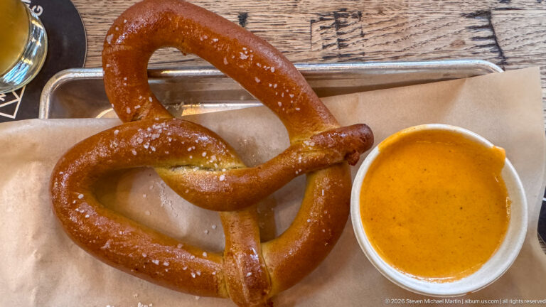 Large salted soft pretzel on brown paper in a metal tray beside a small cup of mustard.