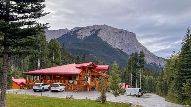 Two-story timber lodge with red metal roof, parked vehicles, forest, and a steep gray mountain behind.