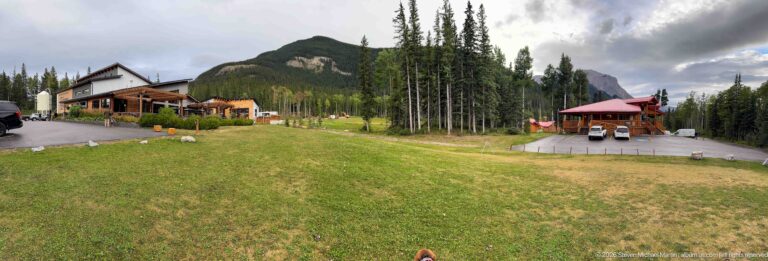 Wide grassy grounds with timber lodge buildings, a red-roofed pavilion, tall conifers, and forested mountains.