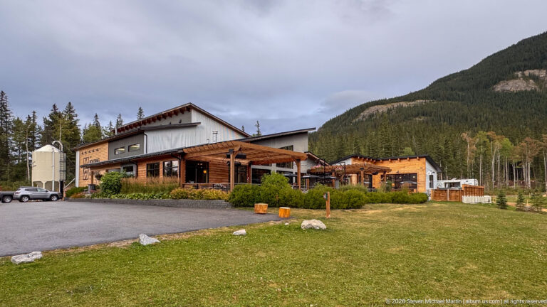 Large timber-accented hospitality building with paved forecourt and grassy verge beneath a forested mountain slope.