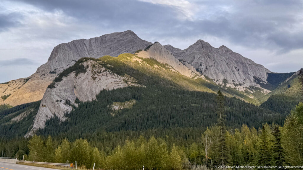Large gray layered mountain with jagged summits and a sunlit lower ridge rising above dense conifer forest.