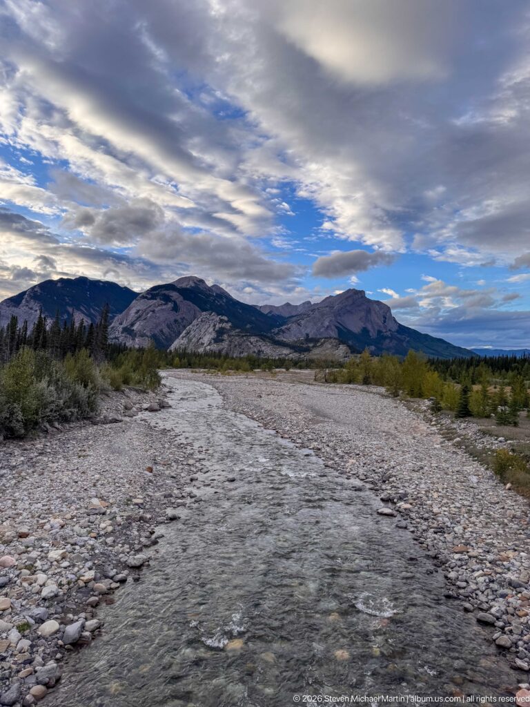 Shallow mountain river flowing through broad pale gravel bars with shrubs, distant ridges, and layered clouds.