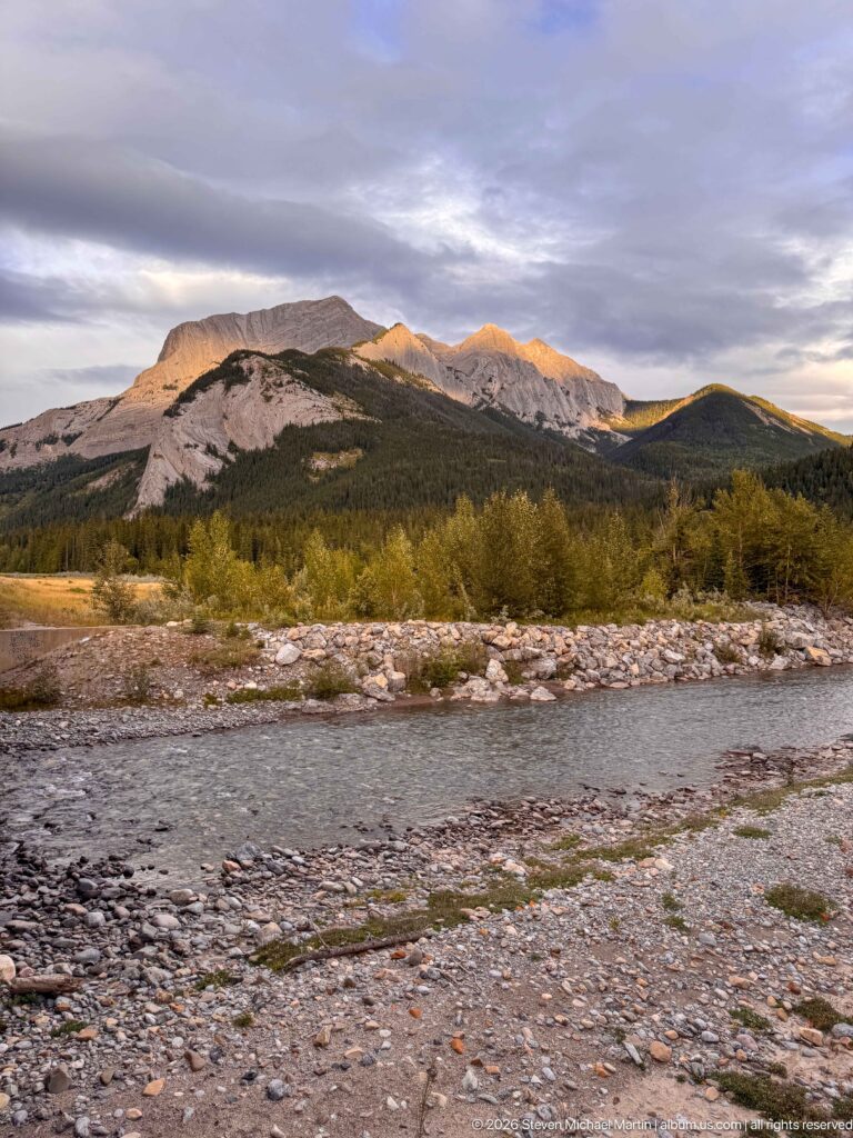 Shallow mountain river crossing a broad gravel floodplain with shrubs, forest, and sunlit ridges in the distance.