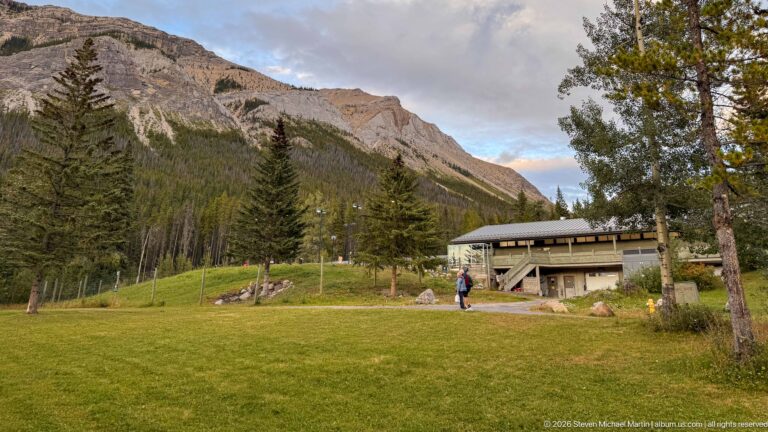 Broad lawn with a few visitors leading to the Miette Hot Springs building beneath forested and rocky mountain slopes.