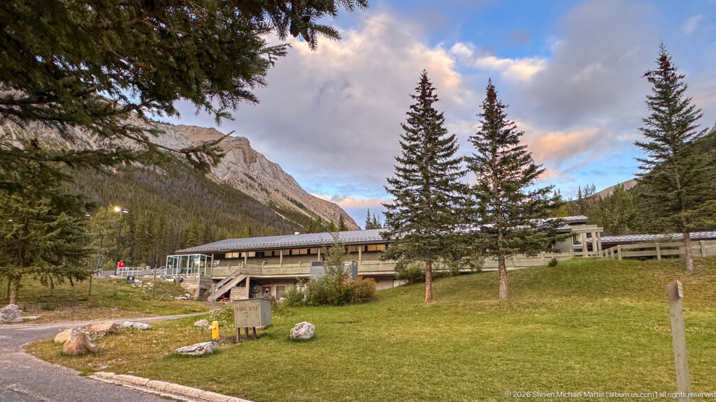 Miette Hot Springs building across a grassy lawn with spruce trees, boulders, paths, and a rocky mountain ridge behind.