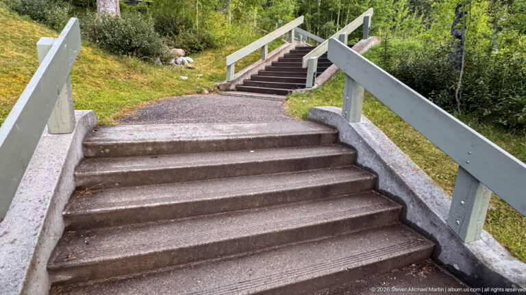 Concrete outdoor staircase with pale railings climbing through grass, gravel paths, shrubs, and boulders.