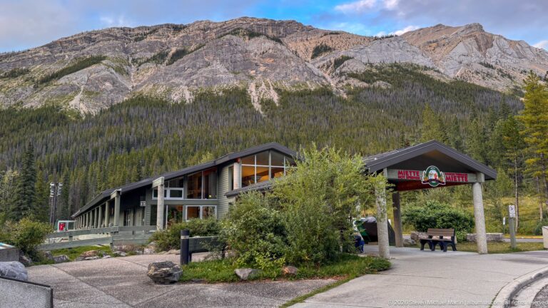 Wide view of the Miette Hot Springs complex, entrance pavilion, paths, shrubs, and long building beneath a massive rocky mountain.