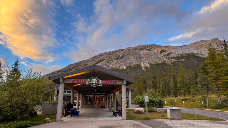 Covered Miette Hot Springs entrance pavilion with signage beneath a steep gray mountain and warm evening clouds.
