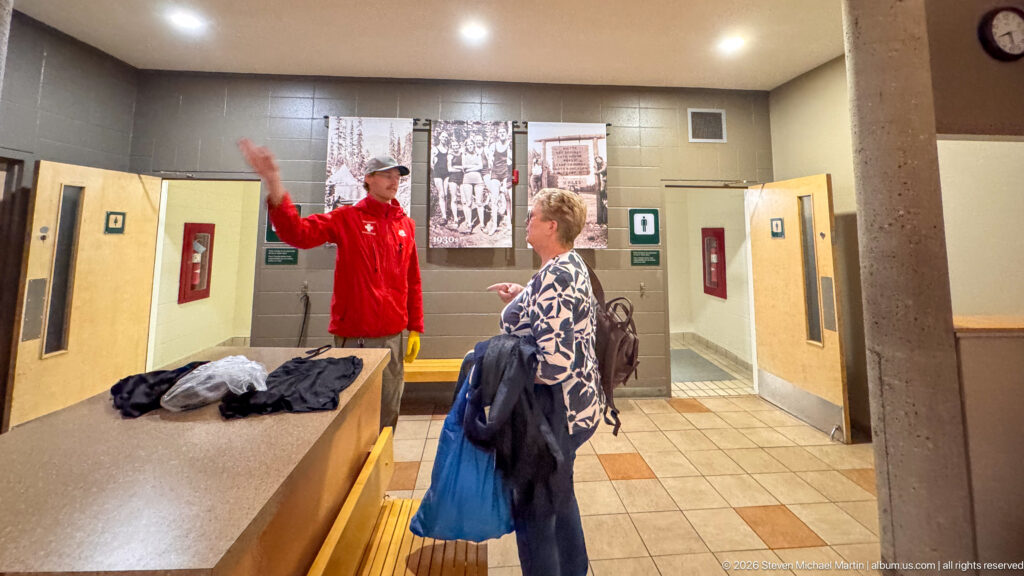 Staff member in red speaking with a visitor inside a tiled changing area with doors and historic photographs.