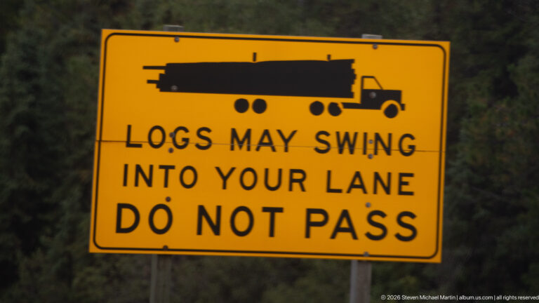 Yellow roadside sign with logging truck silhouette reading Logs may swing into your lane, Do not pass.