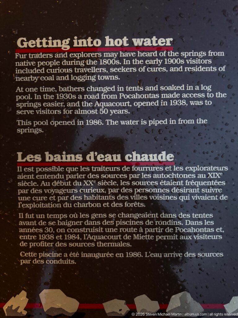Rain-speckled bilingual interpretive panel titled into hot water and Les bains d’eau chaude with historical text.
