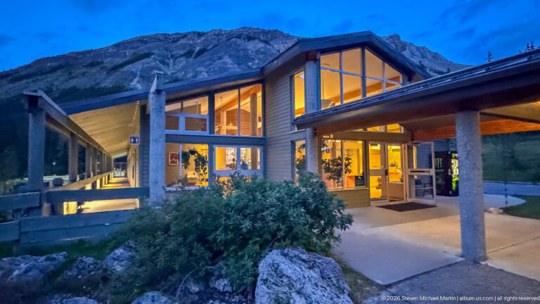 Peaked glass entrance to Miette Hot Springs glowing at dusk, with a shrub, boulder, covered walkway, and mountain backdrop.