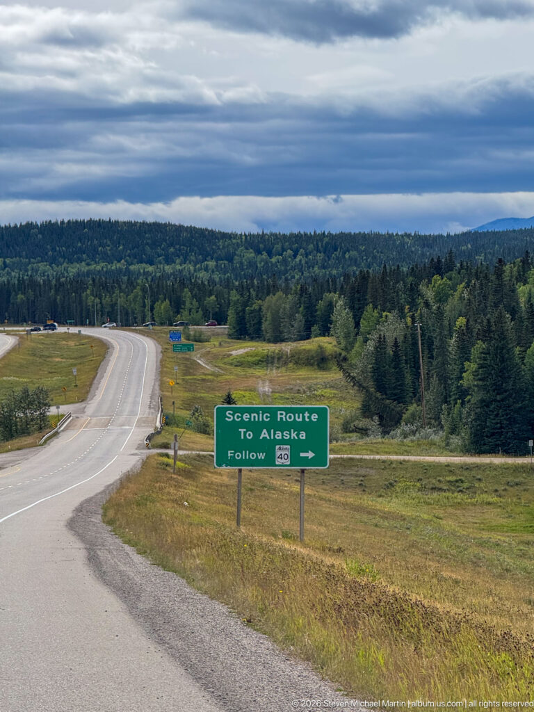 Broad highway through grass and forest with the Scenic Route to Alaska sign beside the junction under cloudy sky.