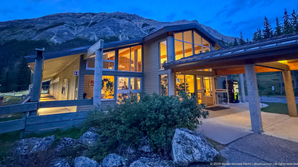 Illuminated peaked entrance of Miette Hot Springs at dusk with concrete columns, shrubs, and a steep mountain behind.