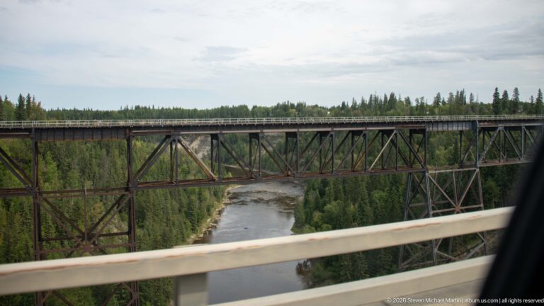 Tall steel trestle bridge with triangular bracing spanning a river and forested valley.