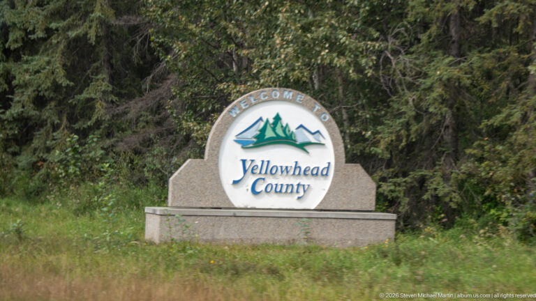 Stone roadside monument with a circular Welcome to Yellowhead County emblem against dense green forest.