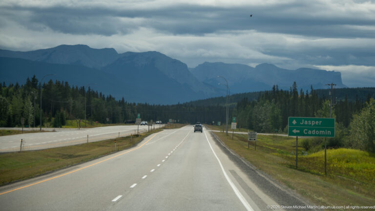 Highway through forested mountains with a green sign pointing ahead to Jasper and left to Cadomin.