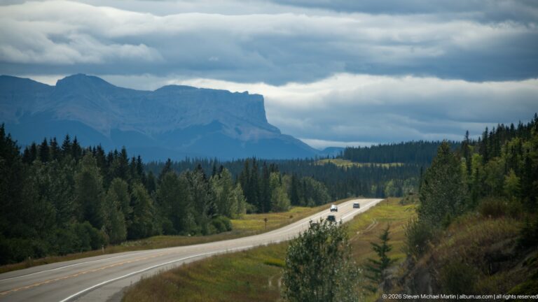Highway with several vehicles crossing open grass and forest toward a broad flat-topped mountain under storm clouds.