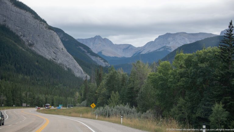 Paved highway curving through a forested mountain valley toward distant jagged peaks beneath cloudy sky.
