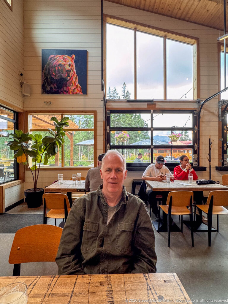 Seated diner at a rustic wood table with large windows, other patrons, and a colorful bear painting behind.